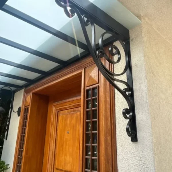 Close-up of a decorative black wrought iron canopy bracket with elegant scrollwork, supporting a frosted glass entrance roof against a white exterior wall.