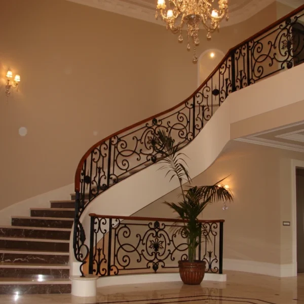 Exquisite curved wrought iron staircase railing with intricate baroque patterns and a dark wood handrail in a grand lobby with a chandelier.