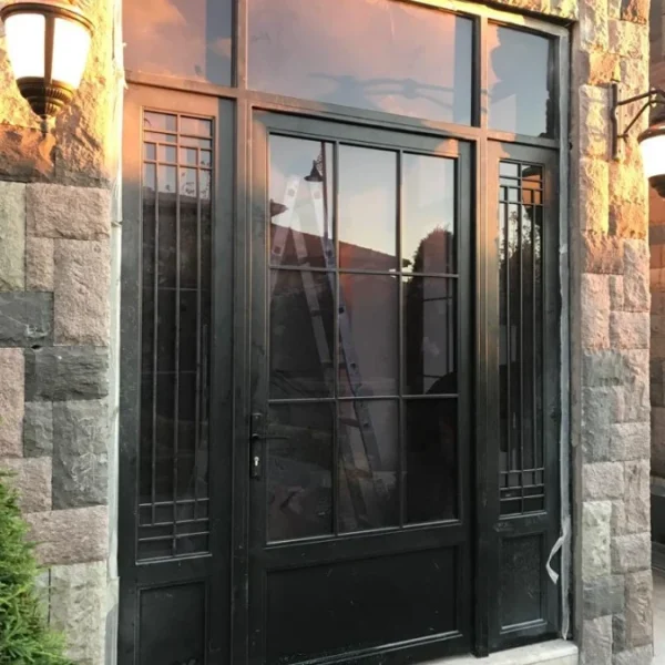 Modern black steel building entrance door with a large glass pane and symmetrical side panels featuring a linear grid pattern, set in a natural stone wall with outdoor lanterns.